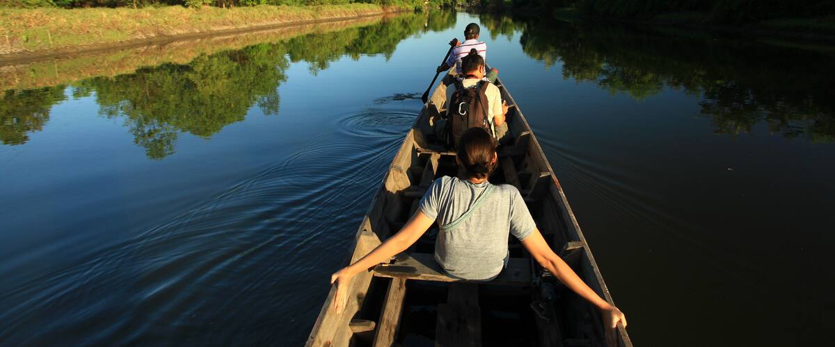 A narrow waterway flanked by dense greenery, with a wooden boat carrying two people.