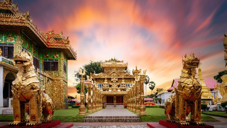 A golden temple with intricate carvings and two large statues, set against a colorful sunset sky.