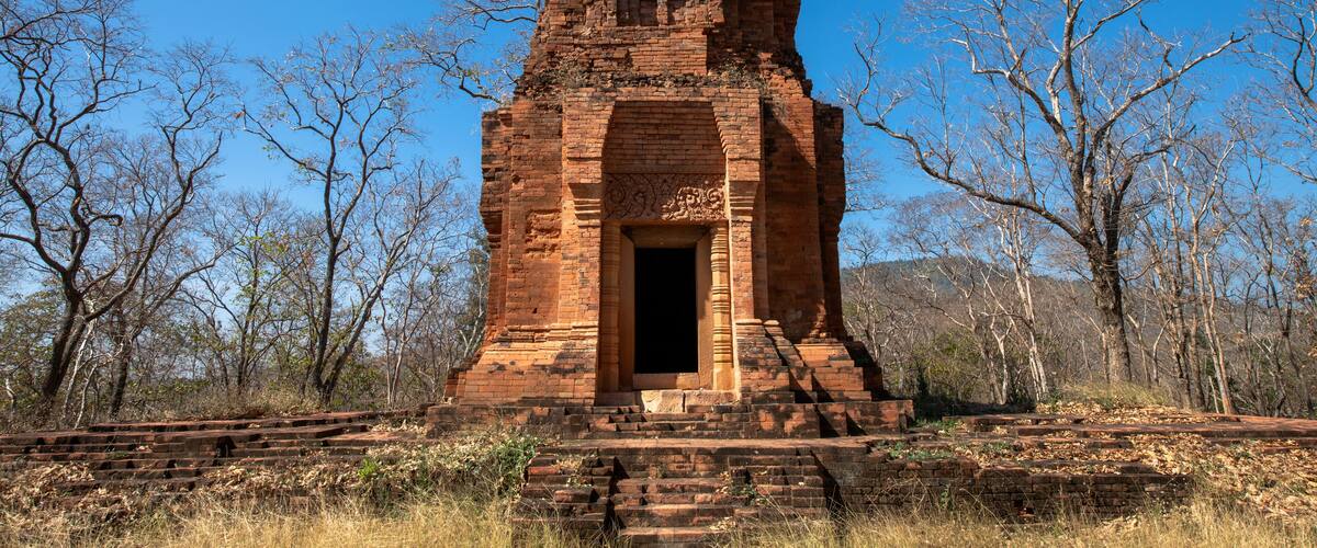 A large, ancient brick structure with a central doorway, surrounded by leafless trees and a clear blue sky.
