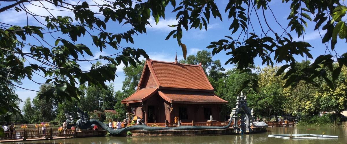 A traditional wooden building with a tiled roof by a calm pond, surrounded by lush greenery and a decorative bridge.