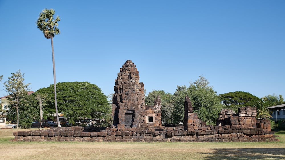 An ancient stone structure with a central tower and smaller buildings, surrounded by trees and grass.