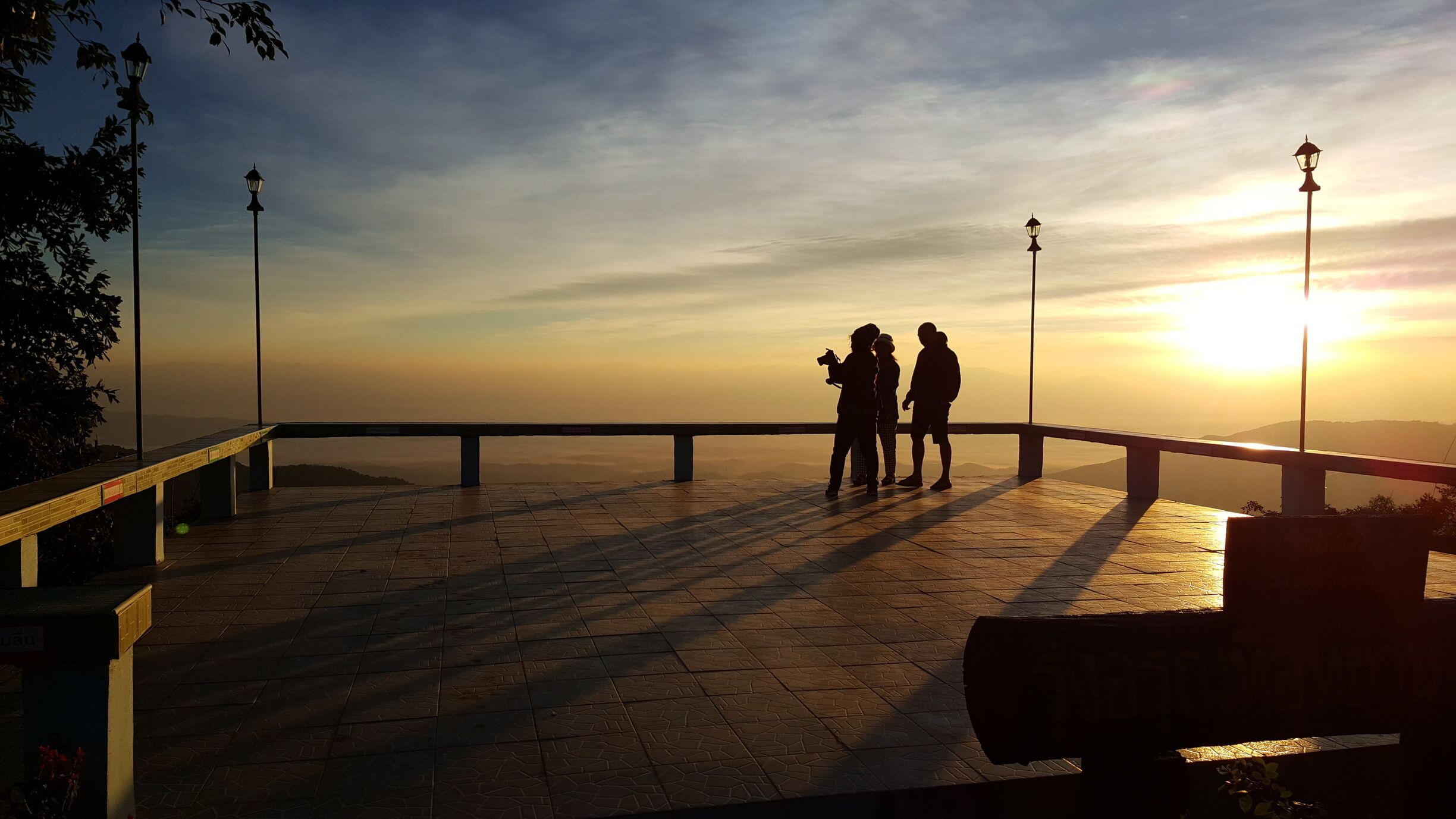 A sunset over a paved viewing platform with railings and street lamps.