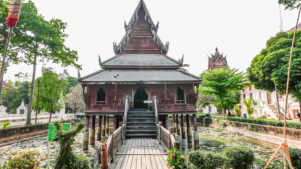A traditional Thai temple with a multi-tiered roof, a wooden staircase leading up to the entrance, and a small bridge over a water canal.