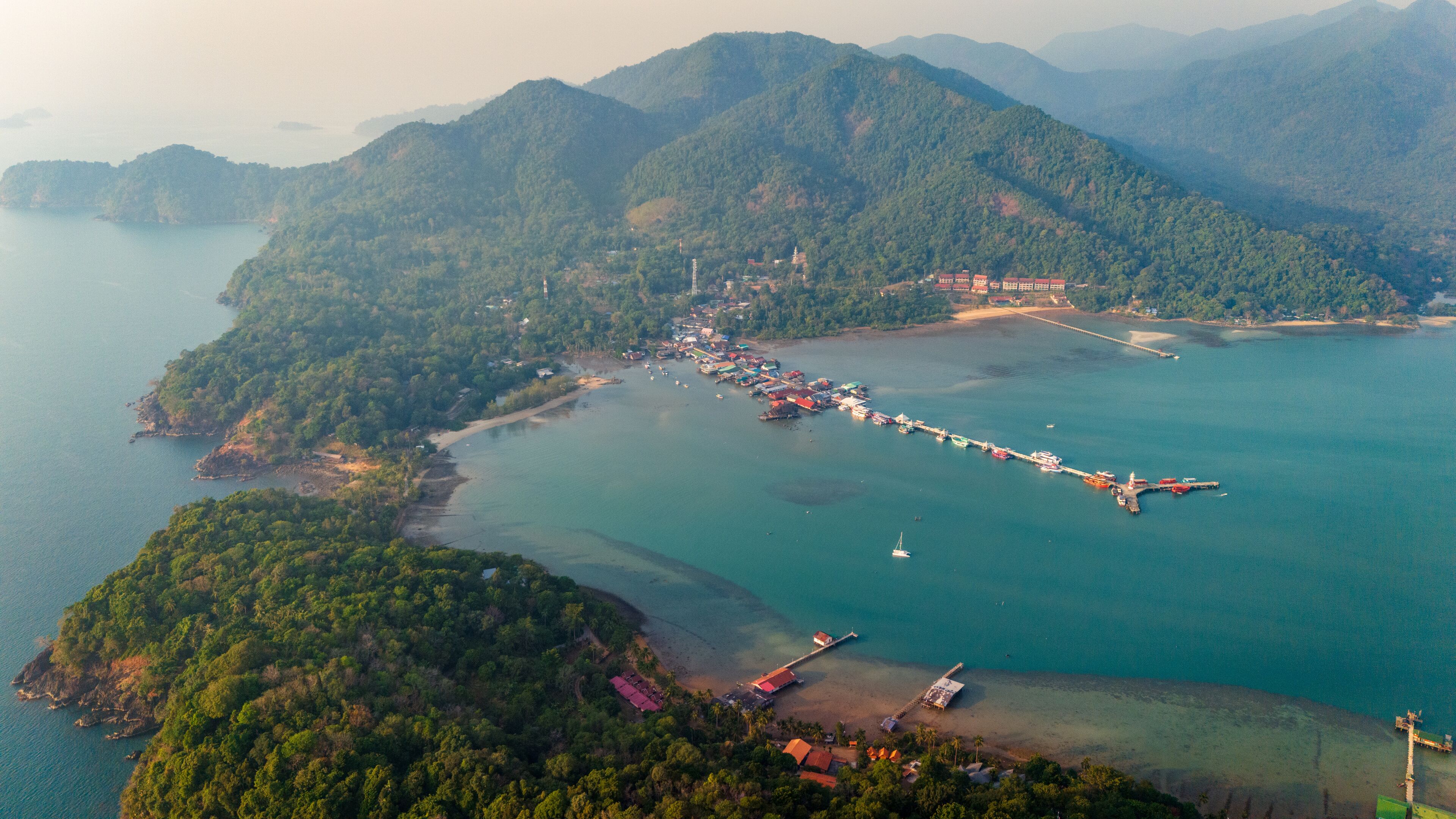 An aerial view of a coastal town with a long pier extending into the sea, surrounded by lush green hills and a clear blue sky.