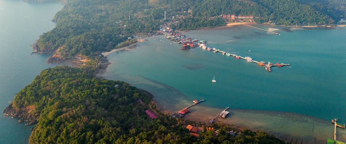An aerial view of a coastal town with a long pier extending into the sea, surrounded by lush green hills and a clear blue sky.
