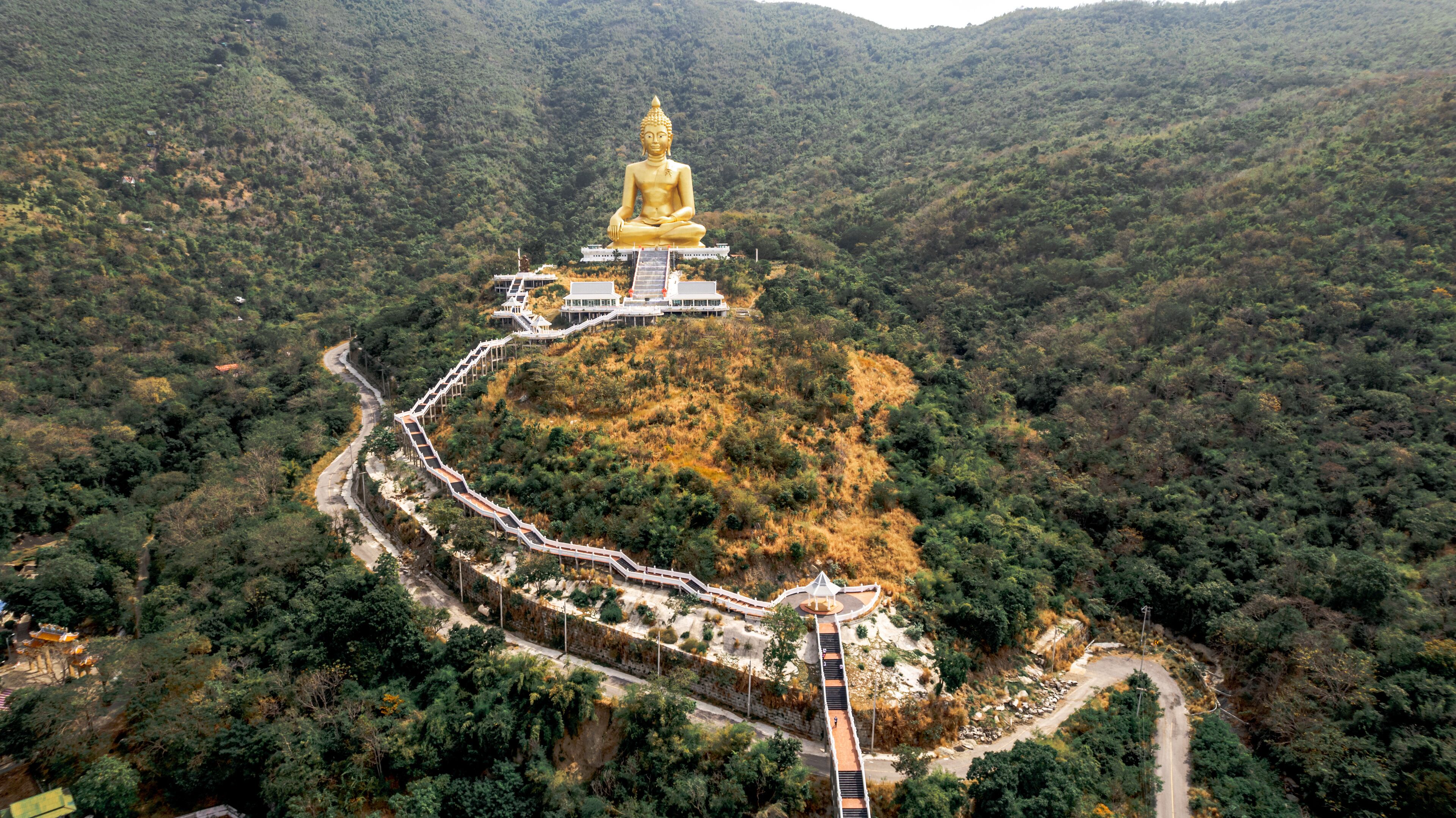 A golden Buddha statue atop a hill, surrounded by lush greenery and a winding path leading to a white building.