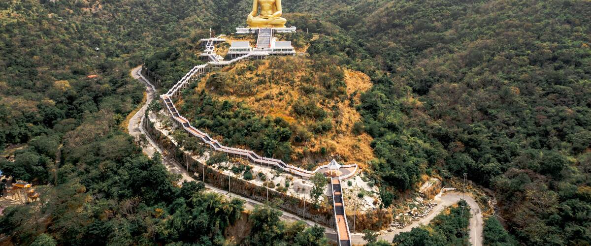 A golden Buddha statue atop a hill, surrounded by lush greenery and a winding path leading to a white building.