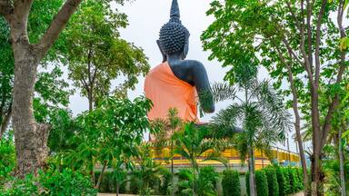 A large Buddha statue in a garden with trees and a yellow building in the background.