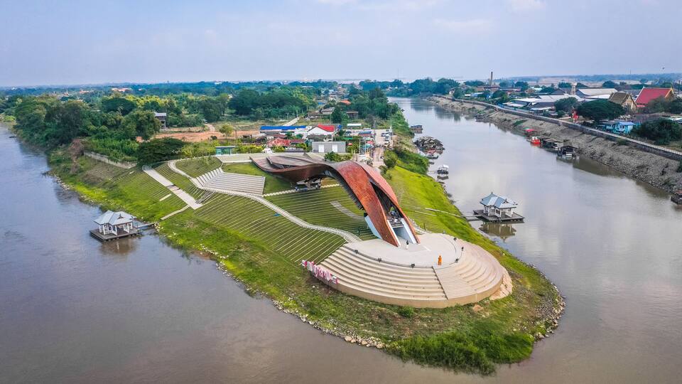 An aerial view of a riverside park with a unique curved bridge, a small boat dock, and a gazebo.