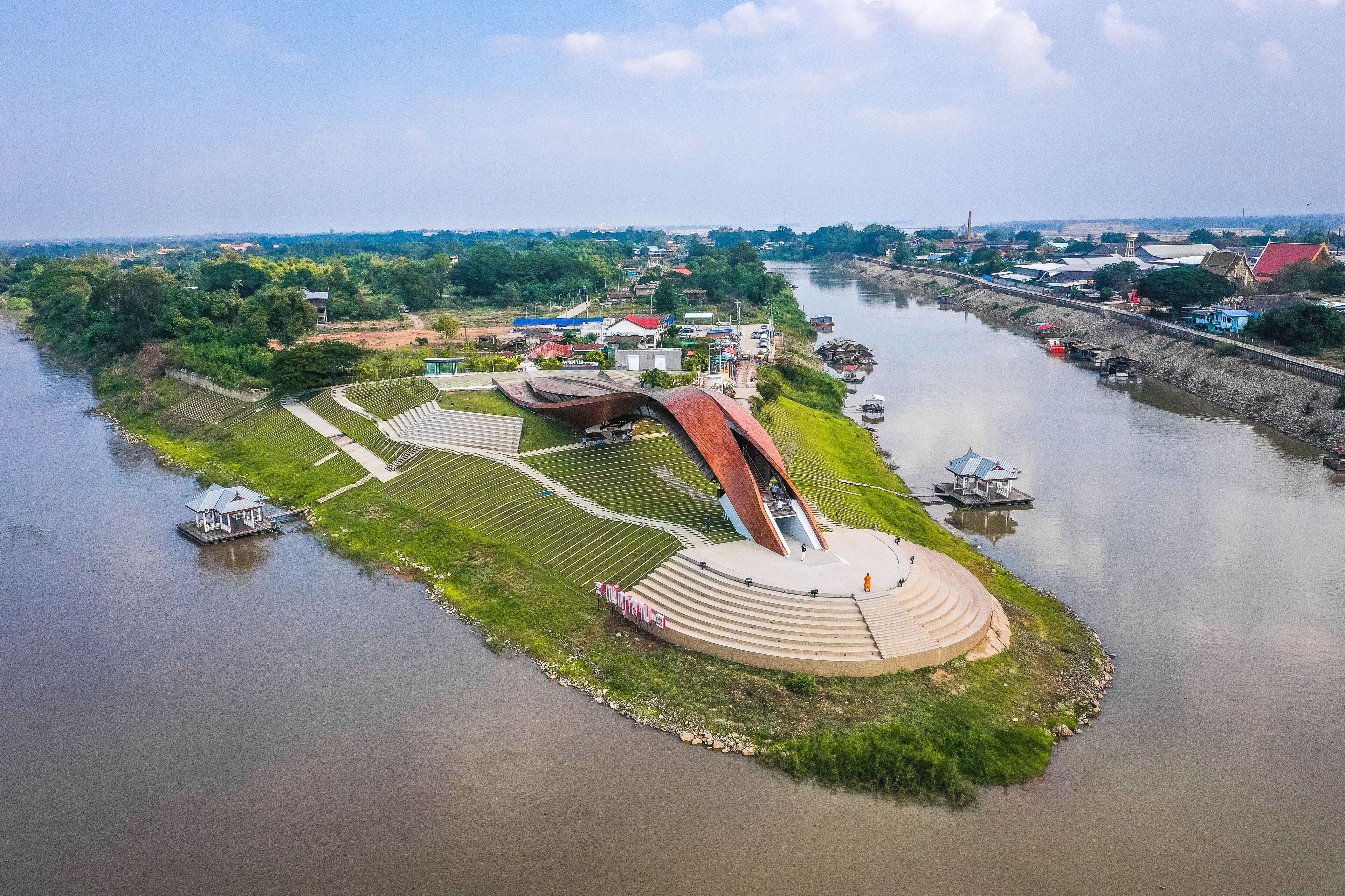 An aerial view of a riverside park with a unique curved bridge, a small boat dock, and a gazebo.