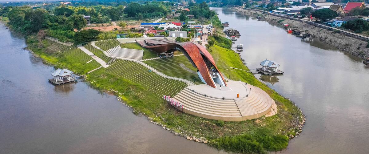 An aerial view of a riverside park with a unique curved bridge, a small boat dock, and a gazebo.