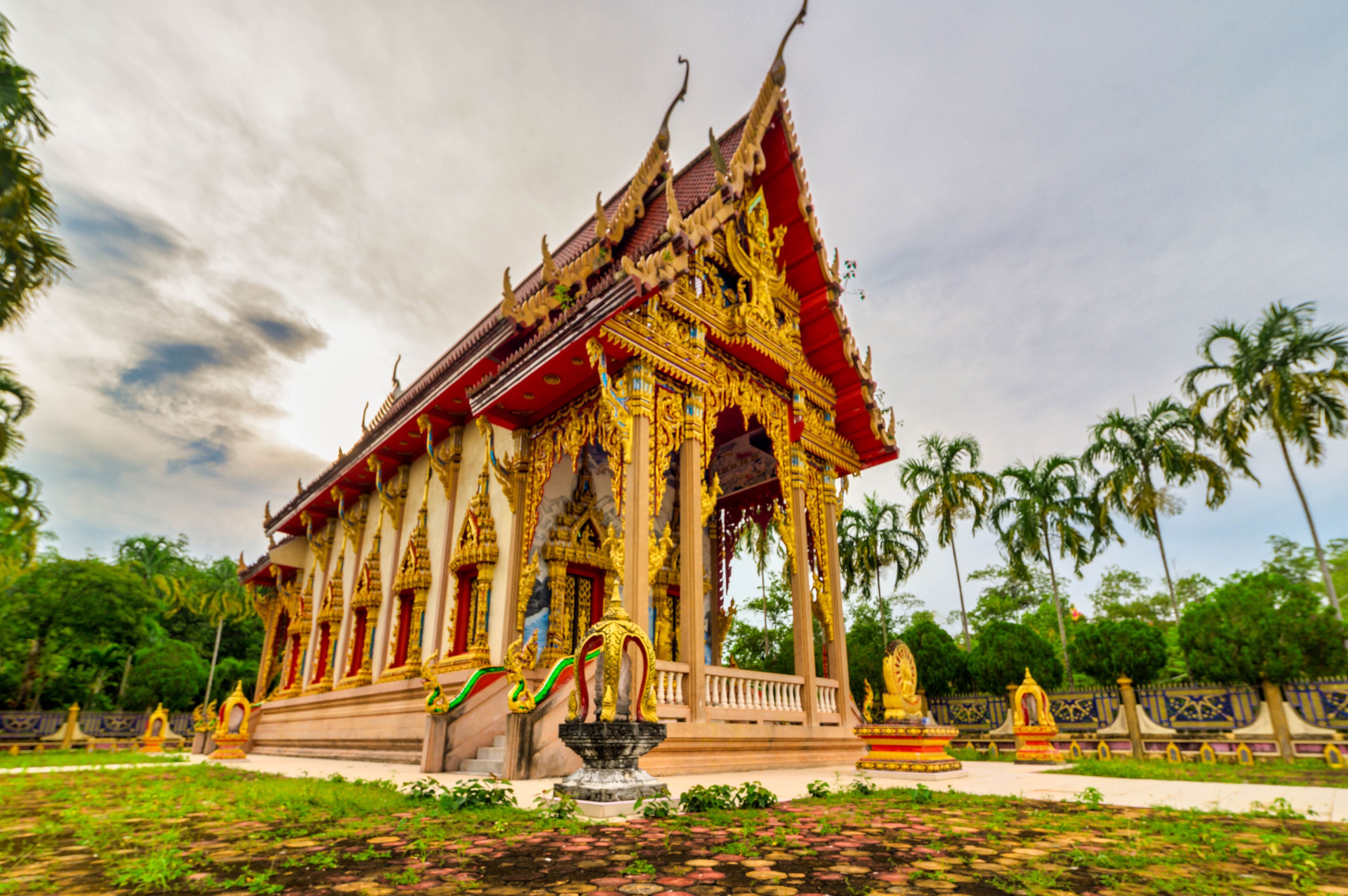 A traditional Thai temple with ornate golden decorations, a central stupa, and a surrounding garden with palm trees.