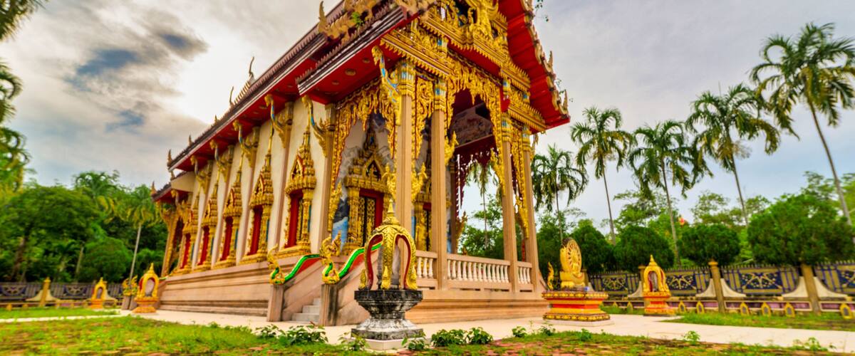 A traditional Thai temple with ornate golden decorations, a central stupa, and a surrounding garden with palm trees.