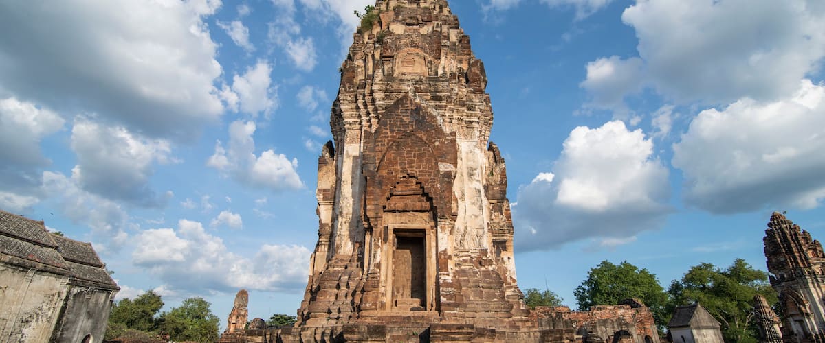 An ancient temple with a central doorway, surrounded by ruins and trees under a partly cloudy sky.