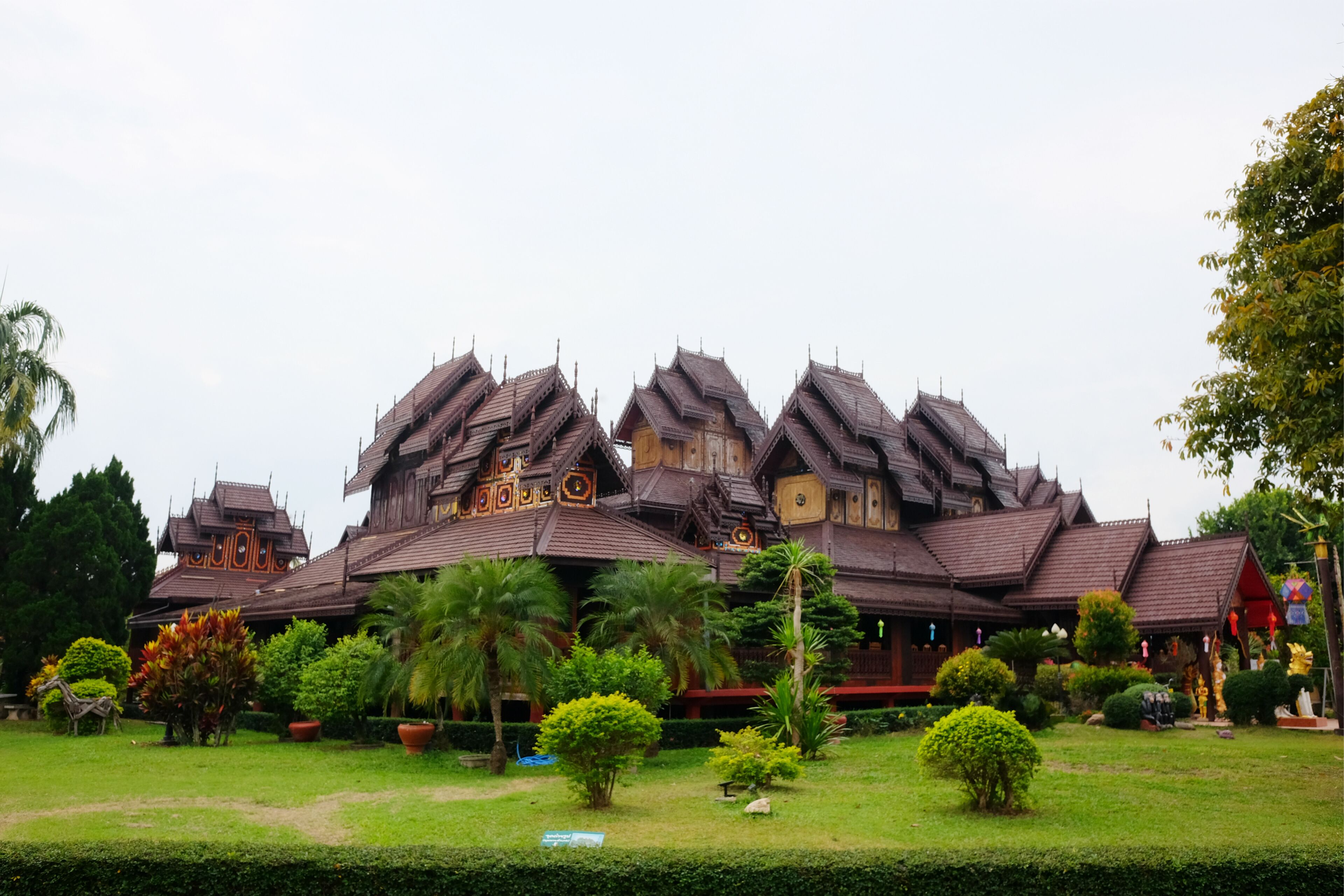 A traditional wooden house with multiple steeply pitched roofs surrounded by well-maintained gardens and palm trees.