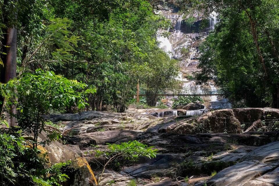 A waterfall cascading over rocks in a forested area with dense greenery and a bridge in the background.