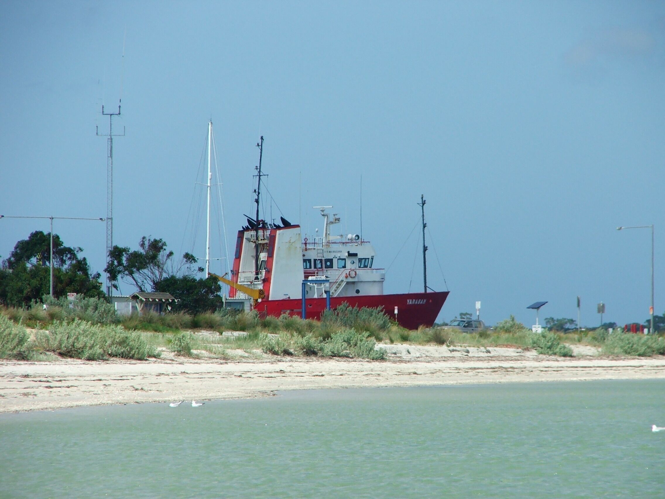 A red and white fishing boat named 'YARADAH' anchored near a sandy beach with sparse vegetation and a clear sky.