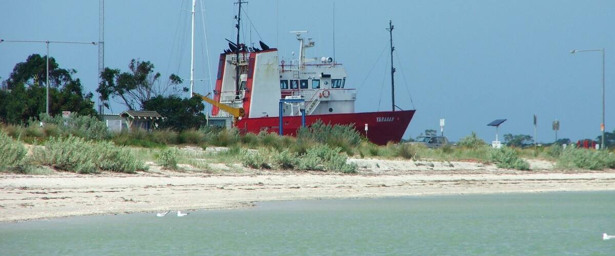 A red and white fishing boat named 'YARADAH' anchored near a sandy beach with sparse vegetation and a clear sky.