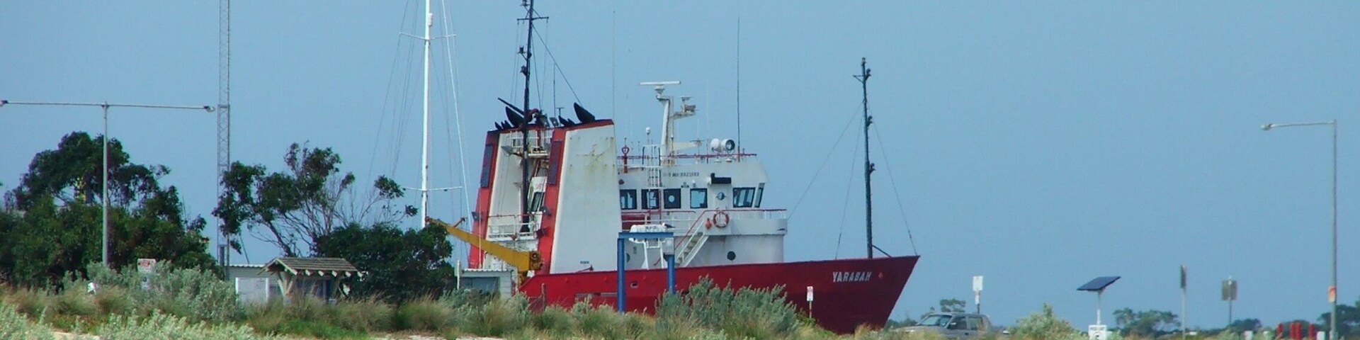 A red and white fishing boat named 'YARADAH' anchored near a sandy beach with sparse vegetation and a clear sky.