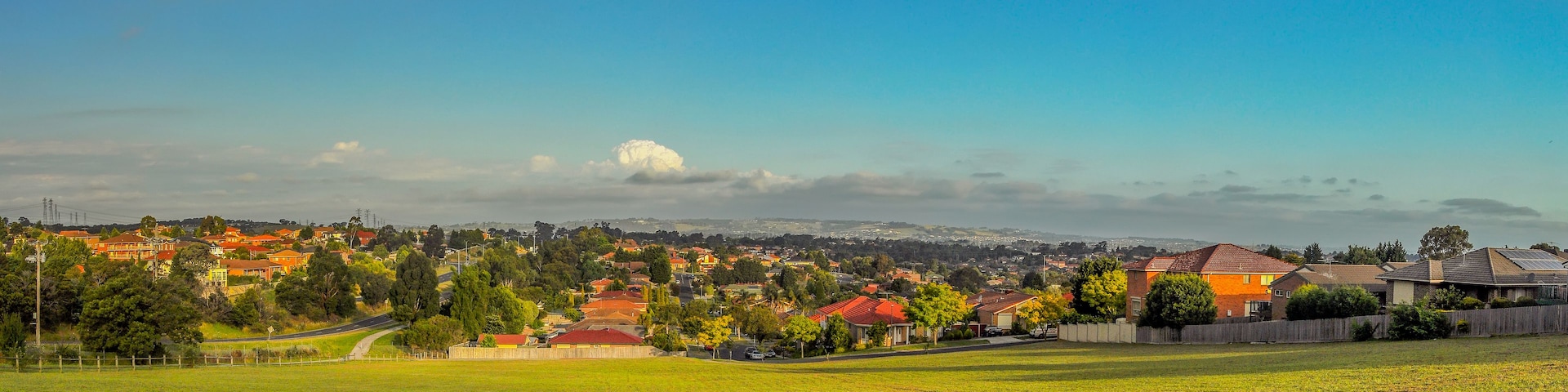 Bairro residencial com casas, um gramado e uma paisagem urbana distante sob um céu claro.