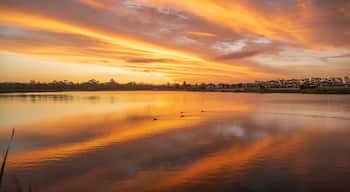Pôr do sol sobre um lago calmo, refletindo o céu, cercado por uma área residencial.