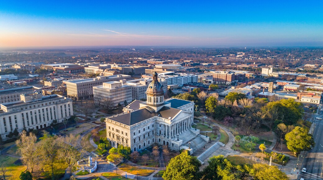 A cityscape with prominent government buildings, including a large white structure with columns and a dome, surrounded by smaller buildings and a mix of green spaces and urban development.