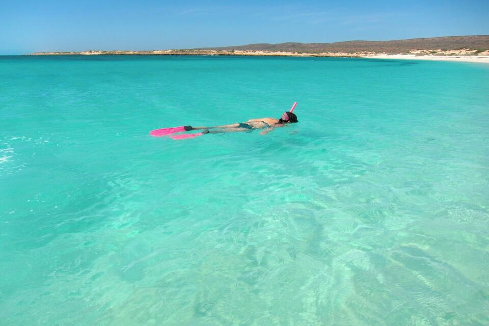 Una persona practica snorkel en aigües turqueses cristal·línes, amb una platja i unes colines al fons.