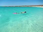 Eine Person schnorchelt in klarem türkisblauem Wasser, im Hintergrund ein Strand und Hügel.