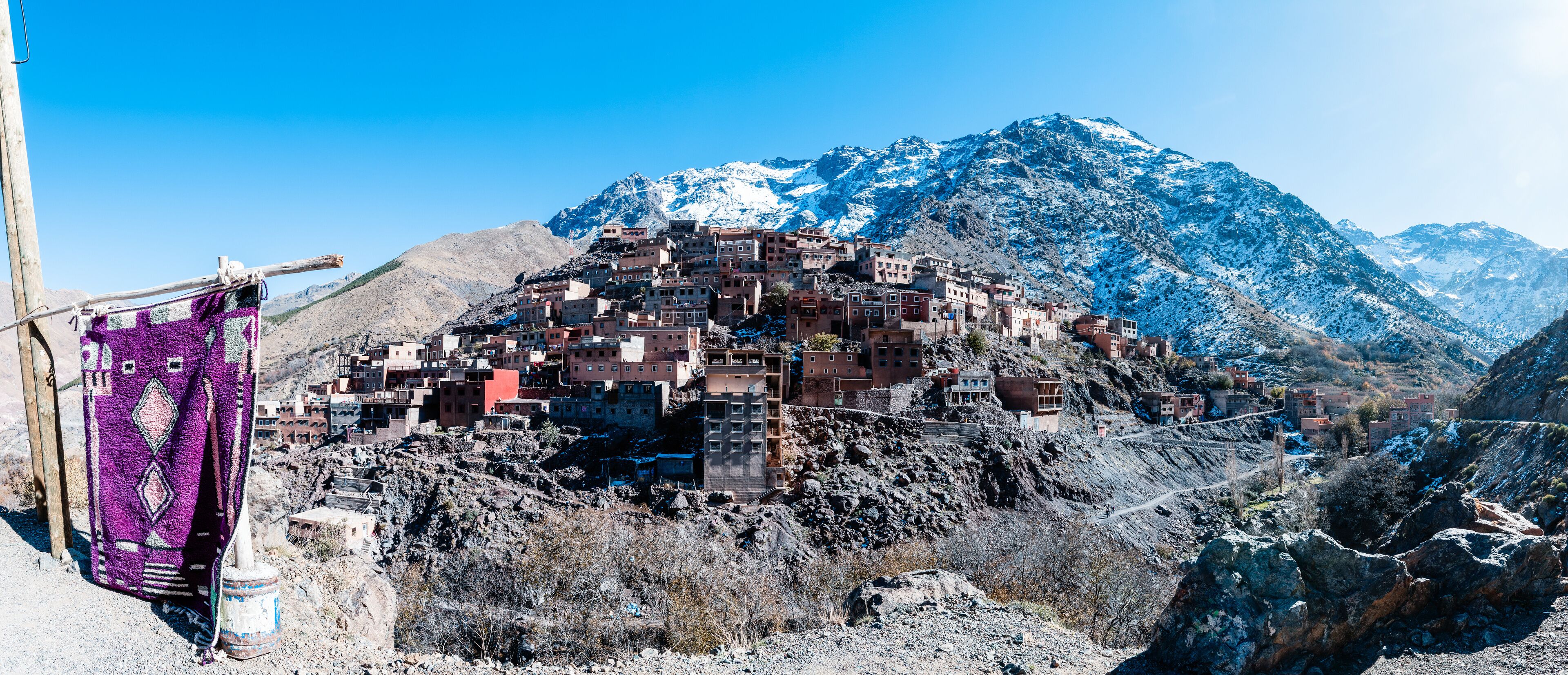 A mountainous village with buildings perched on rocky terrain, a purple cloth hanging on a pole, and snow-capped mountains in the background.