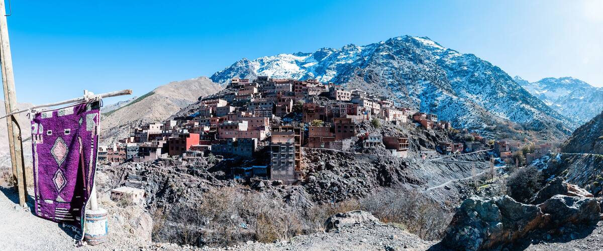 A mountainous village with buildings perched on rocky terrain, a purple cloth hanging on a pole, and snow-capped mountains in the background.