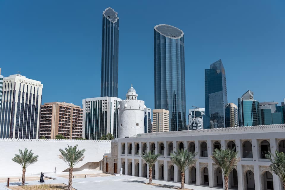 A city skyline with modern skyscrapers, a historic white building with a dome, and palm trees in a courtyard.