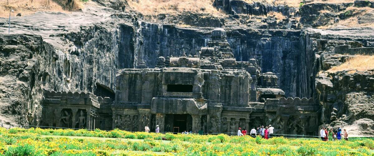 A large rock-cut temple with intricate carvings and a surrounding garden of yellow flowers.