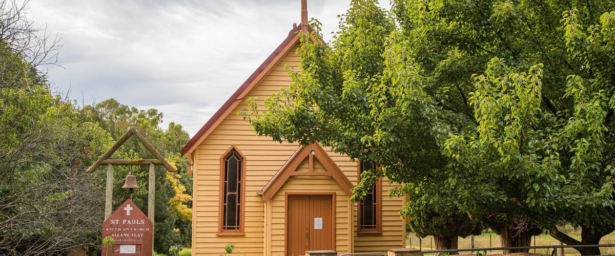 A small wooden church with a cross on top, surrounded by lush greenery and a bell tower.