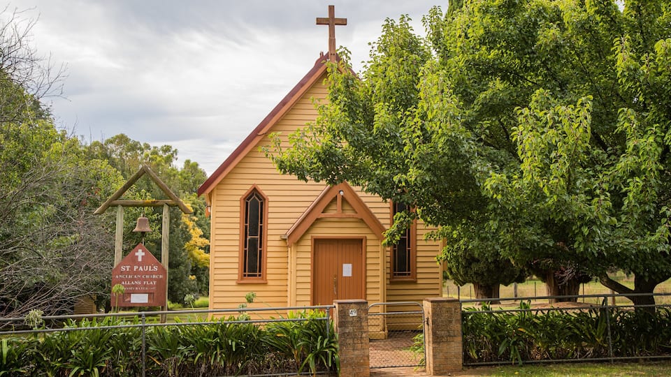 A small wooden church with a cross on top, surrounded by lush greenery and a bell tower.