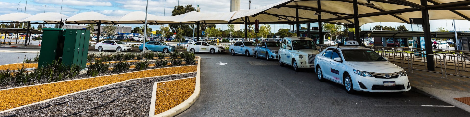An airport terminal with a control tower in the background, covered parking areas, and several taxis parked in front.