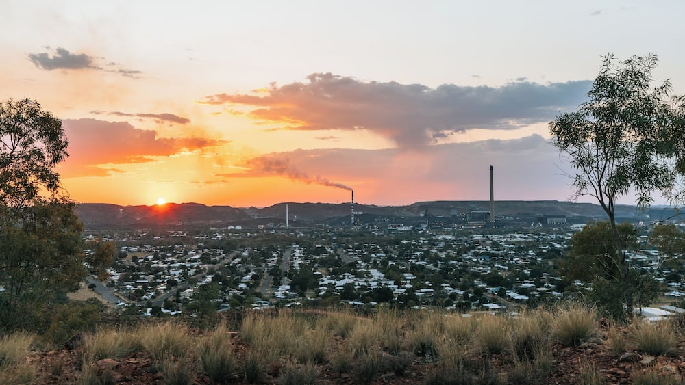 A sunset over a cityscape with industrial chimneys, a river, and surrounding hills.