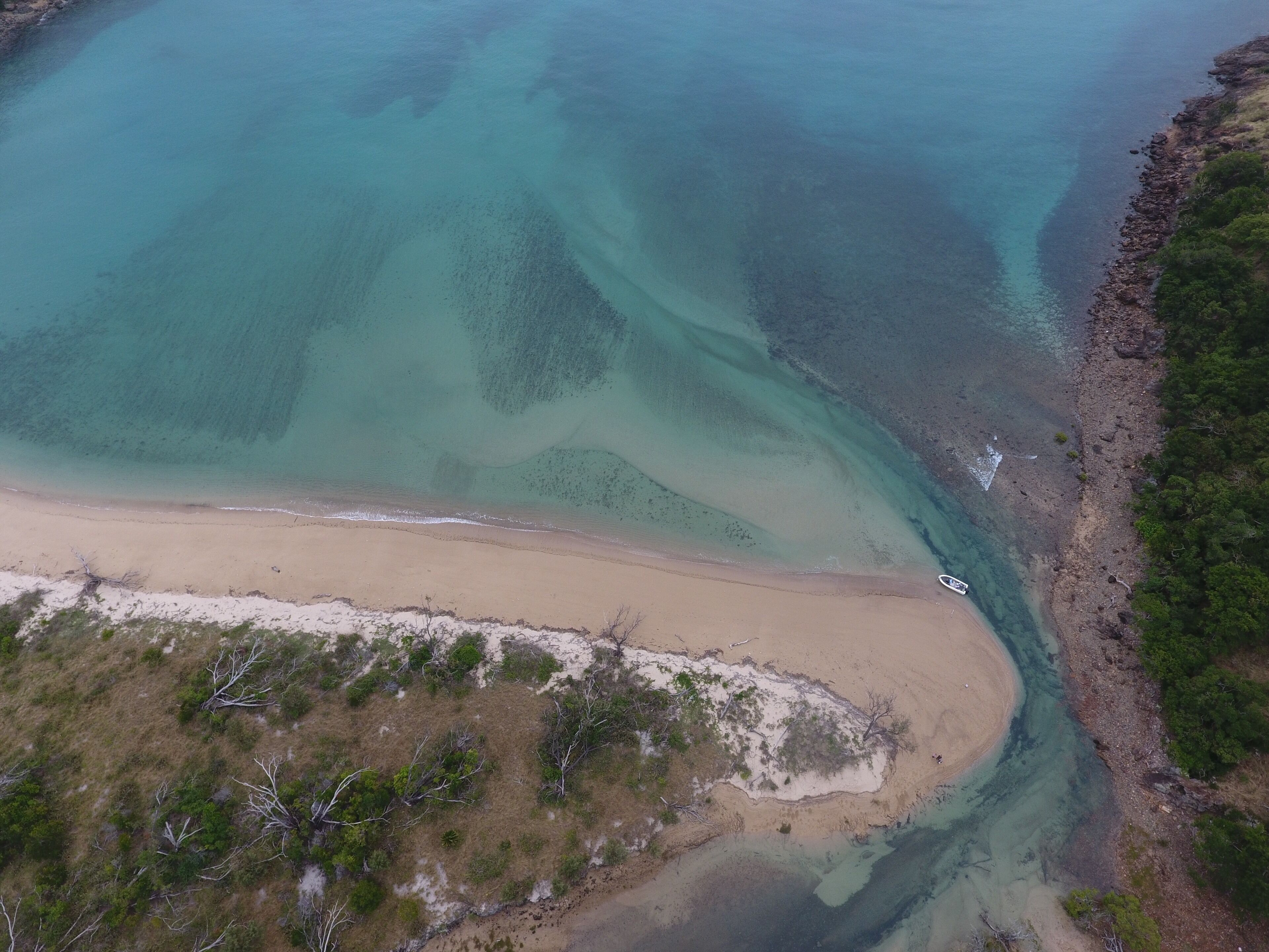 An aerial view of a coastal area with clear turquoise waters, a sandy beach, and a rocky shoreline with sparse vegetation.