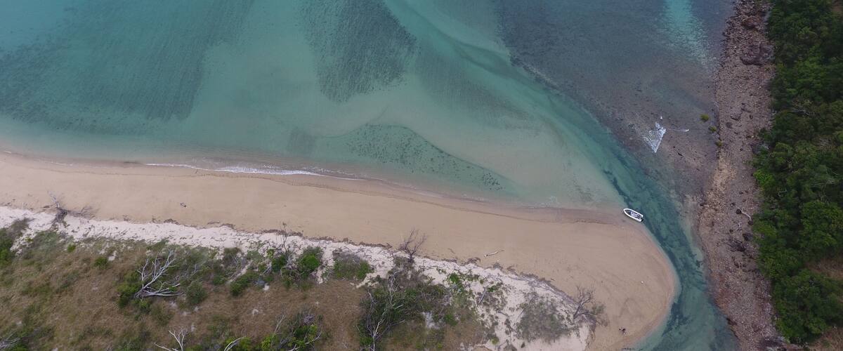 An aerial view of a coastal area with clear turquoise waters, a sandy beach, and a rocky shoreline with sparse vegetation.