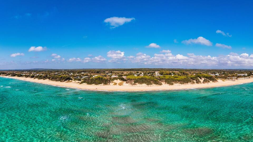 A coastal landscape with clear turquoise waters, a sandy beach, and a small settlement with houses and vegetation.