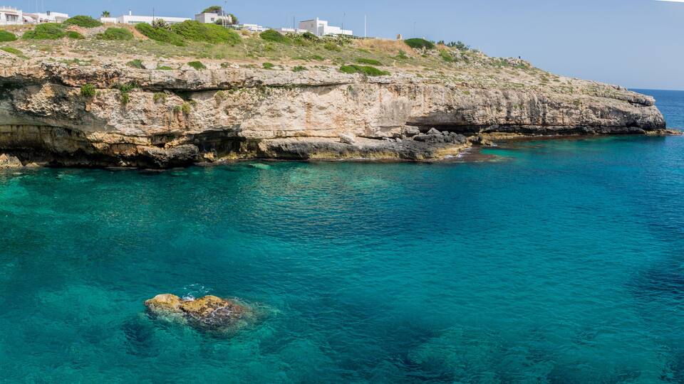 A coastal cliff with clear turquoise water, rocky outcrops, and white buildings on the hillside.
