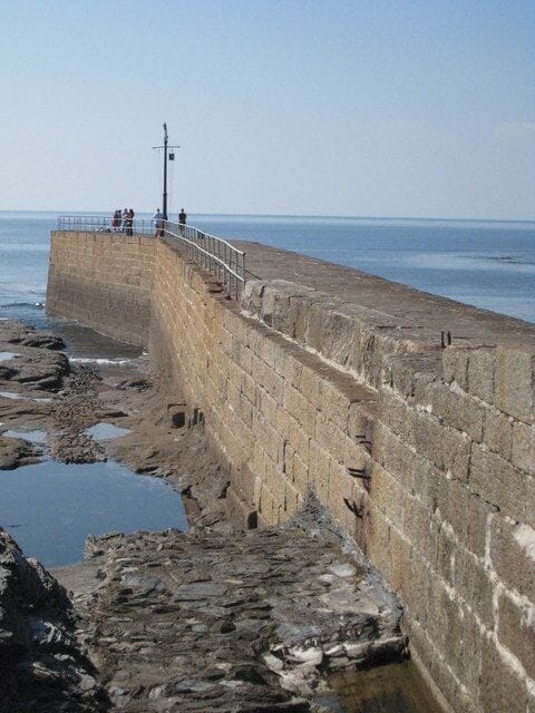 Un muro de piedra junto al mar con una barandilla metálica y un faro al fondo.
