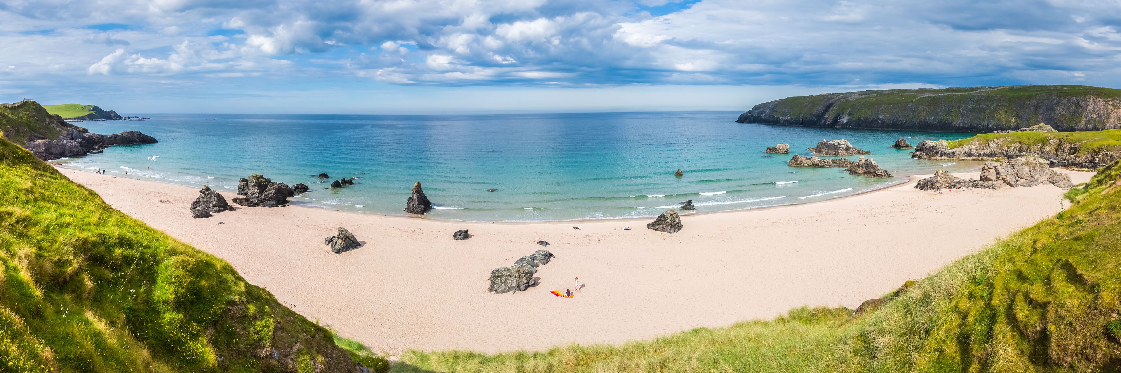 A coastal beach with clear turquoise waters, rocky outcrops, and grassy hills.