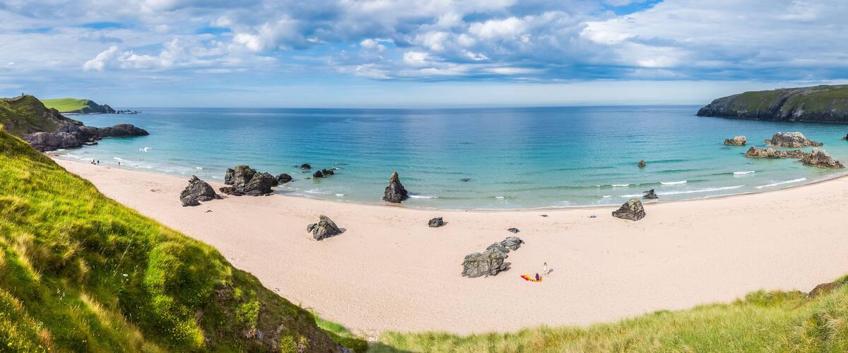 A coastal beach with clear turquoise waters, rocky outcrops, and grassy hills.