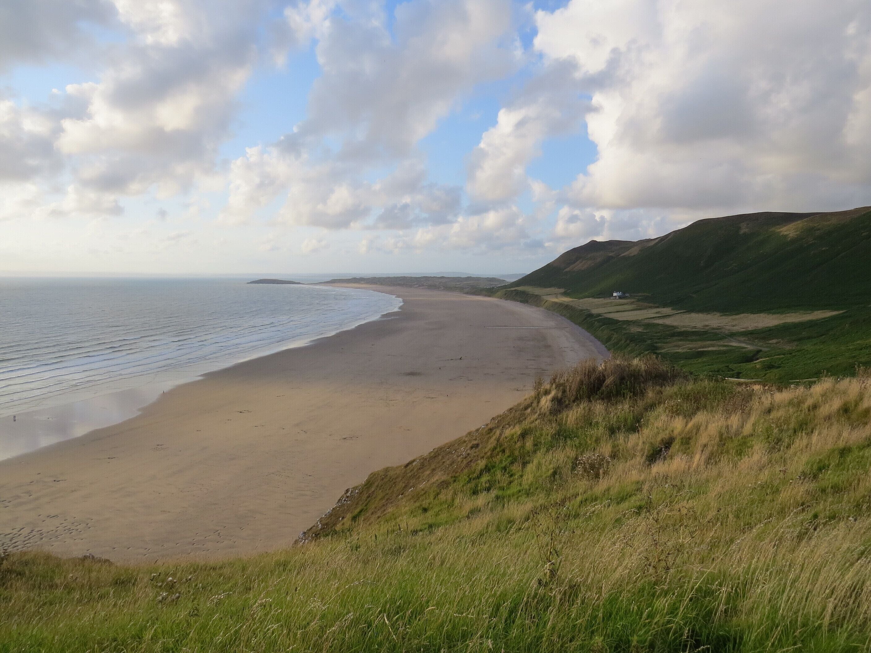 A coastal landscape with a sandy beach, grassy hills, and a partly cloudy sky.
