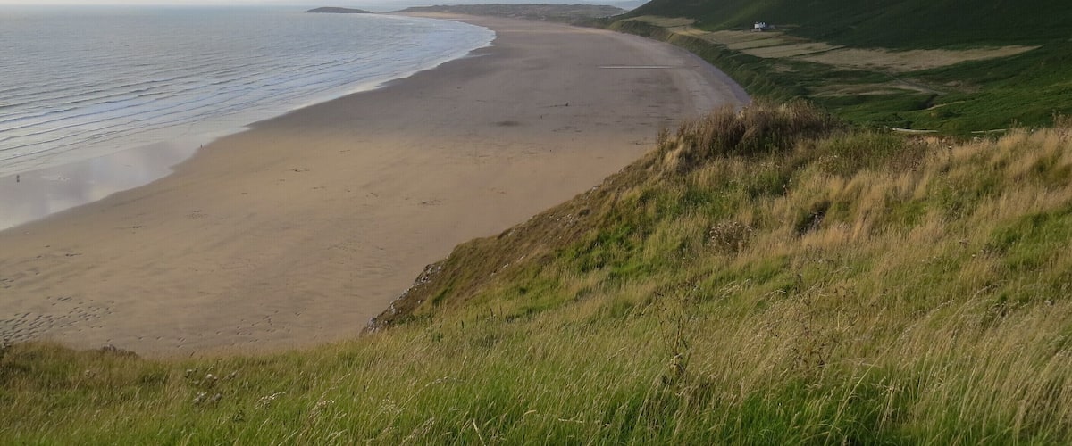 A coastal landscape with a sandy beach, grassy hills, and a partly cloudy sky.