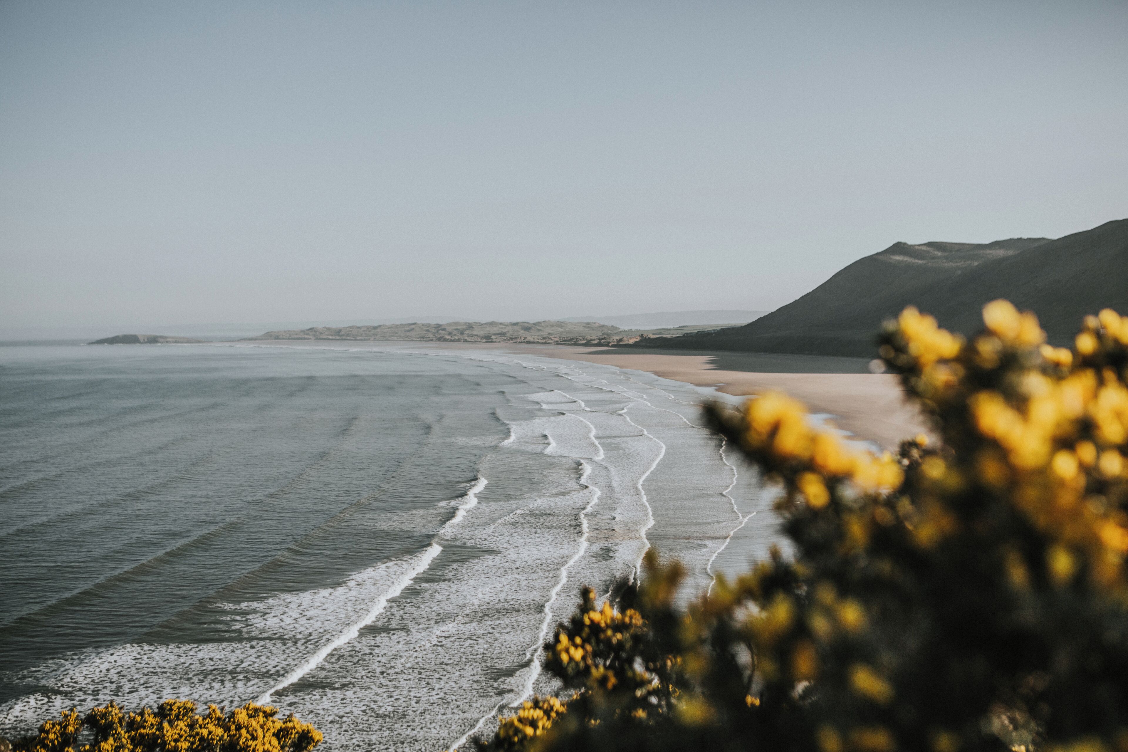 A coastal landscape with a sandy beach, gentle waves, and distant mountains.