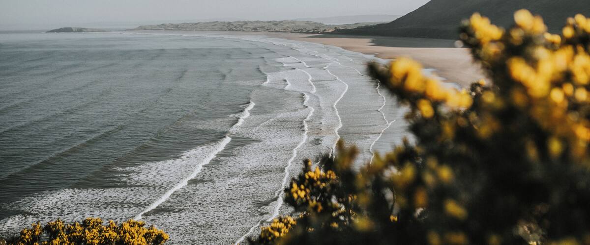 A coastal landscape with a sandy beach, gentle waves, and distant mountains.