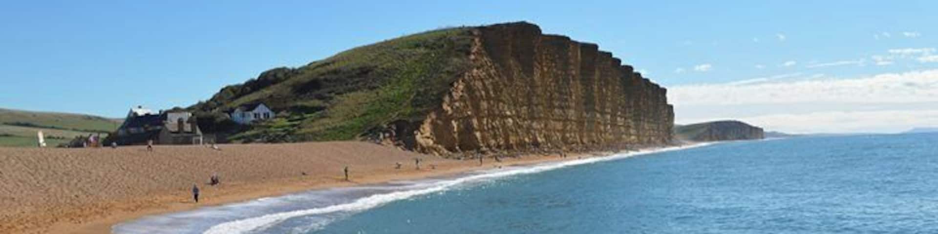 A coastal beach with cliffs, clear blue water, and a few people walking along the shore.