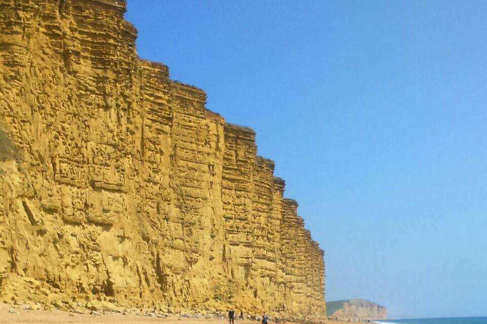 A coastal cliff with layered rock formations and a sandy beach.