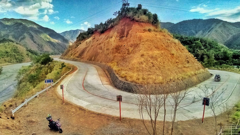 A winding road through a mountainous landscape with a dirt path and a motorcycle parked on the side.